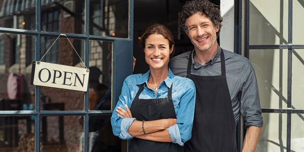 Smiling man and woman in aprons stand at the entrance of their open shop.