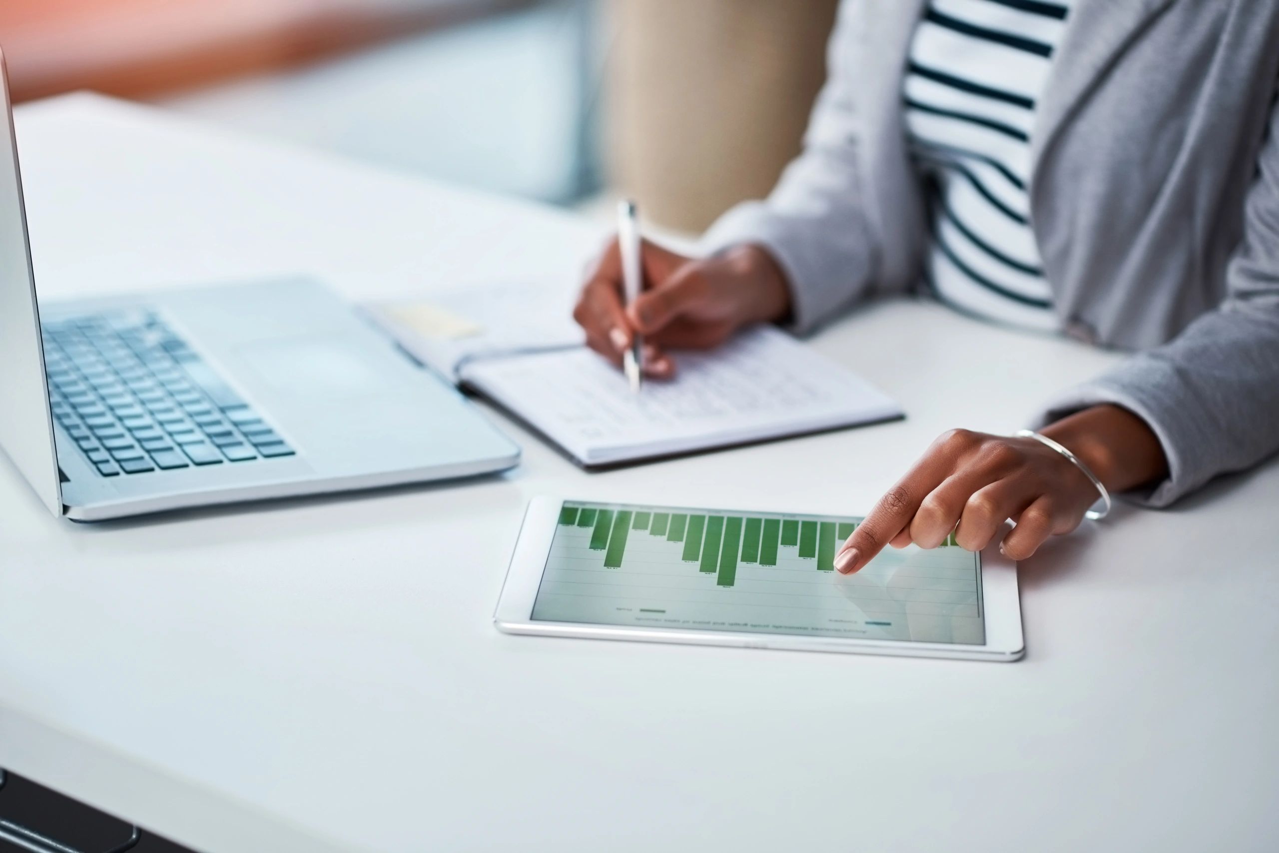 Person analyzing data on a tablet while taking notes beside a laptop.