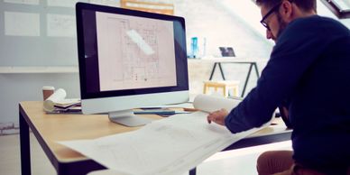 A man reviewing architectural blueprints at a desk with a computer.