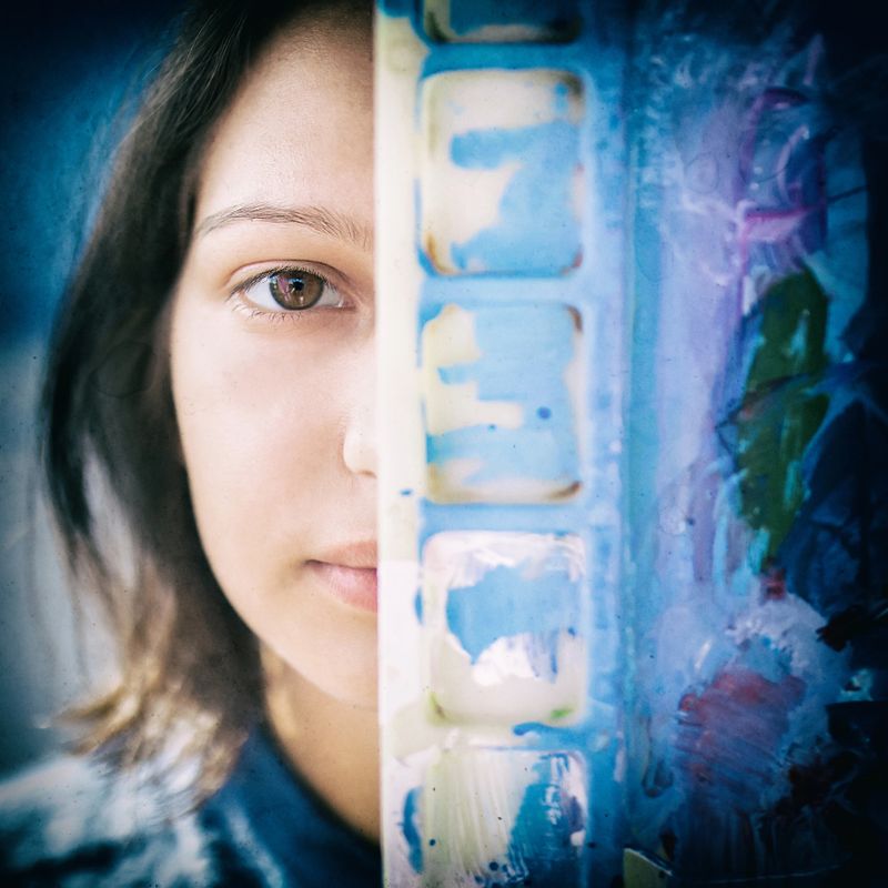 Close-up portrait of beautiful girl artist looking from behind of palette with blue acrylic or oil paint, concept of art.