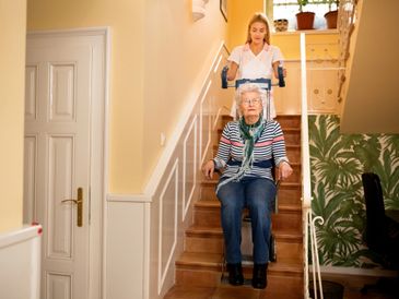 Caregiver assisting elderly woman down stairs with a stairlift.