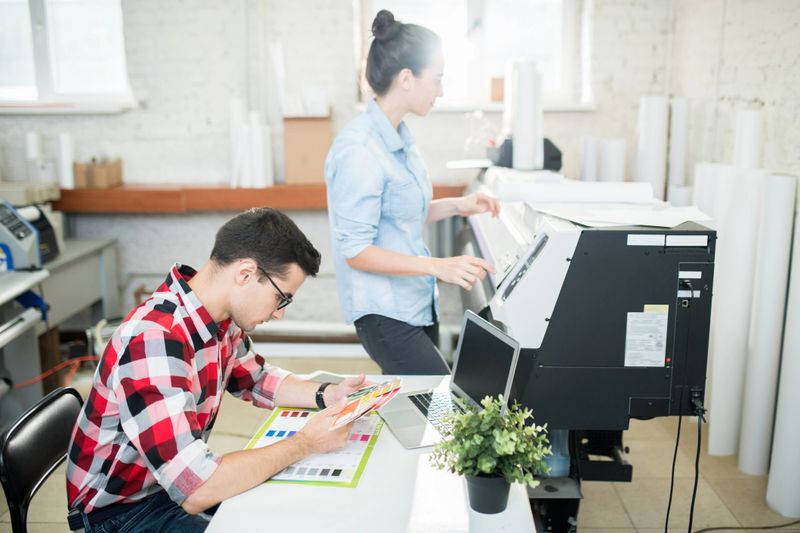 Side view of man exploring palette of colors and woman working with printing machine in modern office