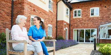 The caregiver is sitting with an older lady outside the assisted living community on a bench.  