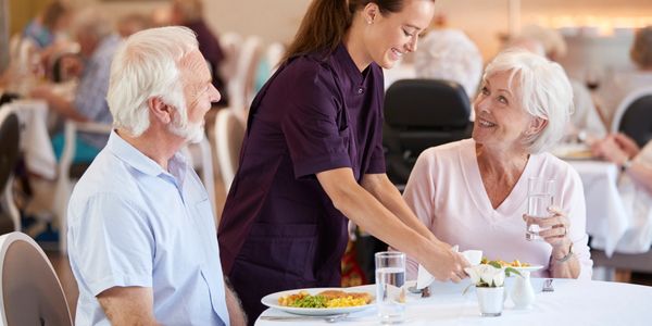 Caregiver serving food to elderly couple at a dining table with smiles.