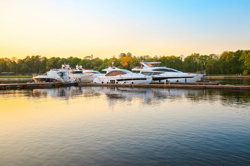 Luxury yachts docked in marina at colorful sunset. Modern motor boats parking at pier. Summer view of river bank, superyachts and water reflection in sunshine. Sunny evening sky. Travel and fashionable vacation.