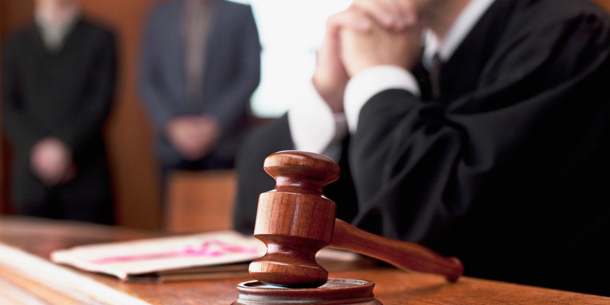 Judge's gavel on desk with blurred people in courtroom background.