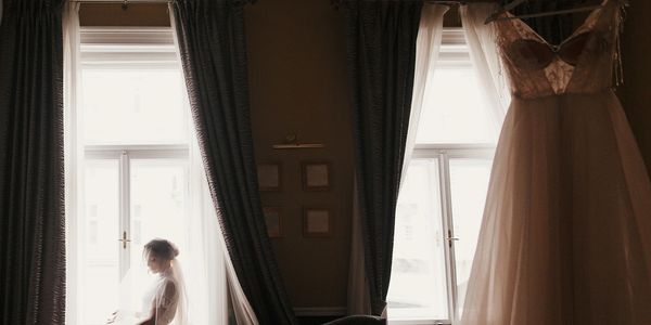 A bride stands by a window with her wedding dress hanging nearby.