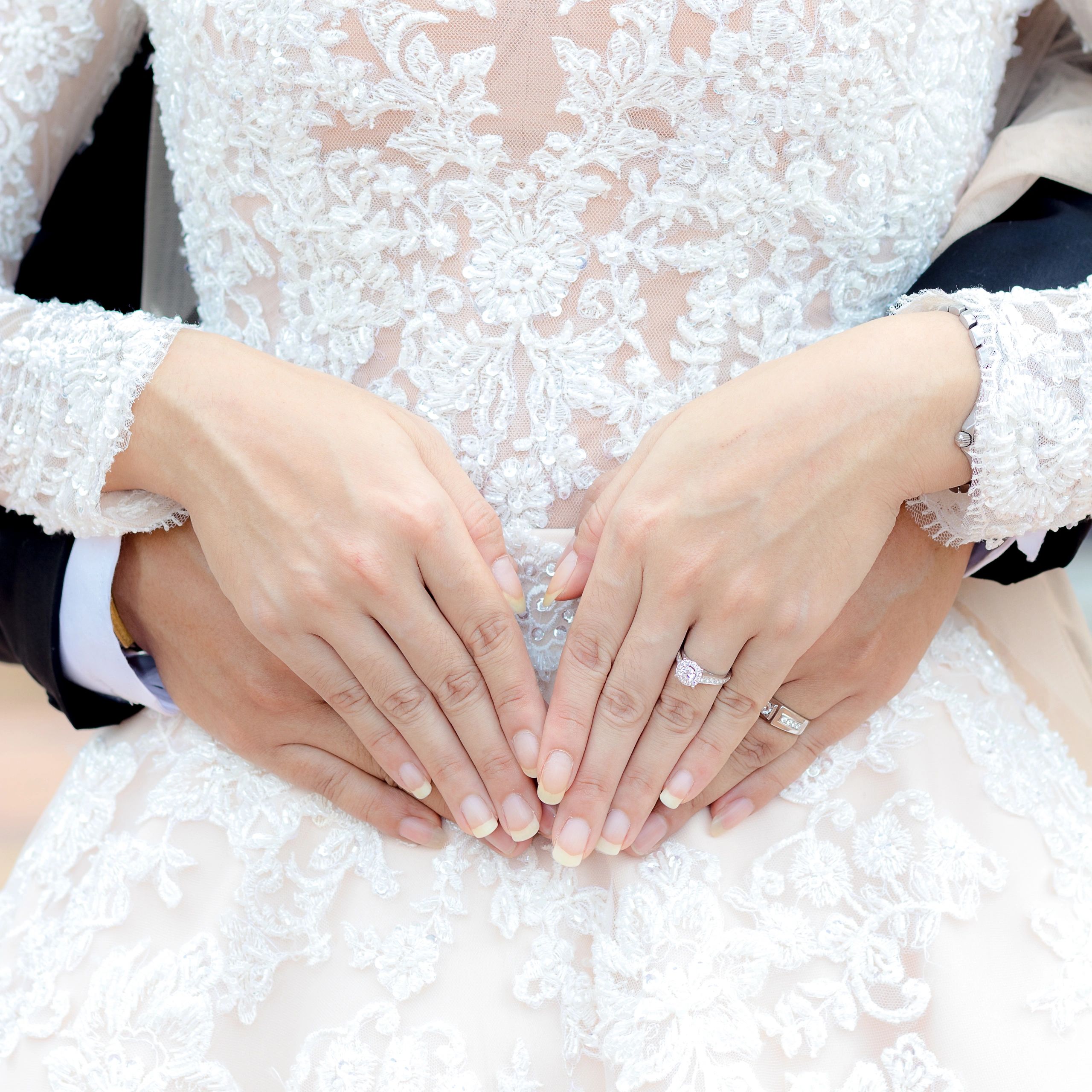 Couple's hands forming a heart on a wedding dress, showcasing wedding rings.