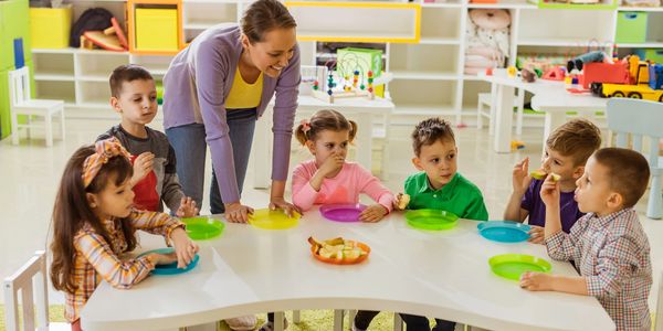 Teacher engaging with children eating snacks at a classroom table.