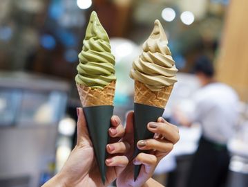 Hands holding matcha and coffee-flavored soft serve ice cream in waffle cones.