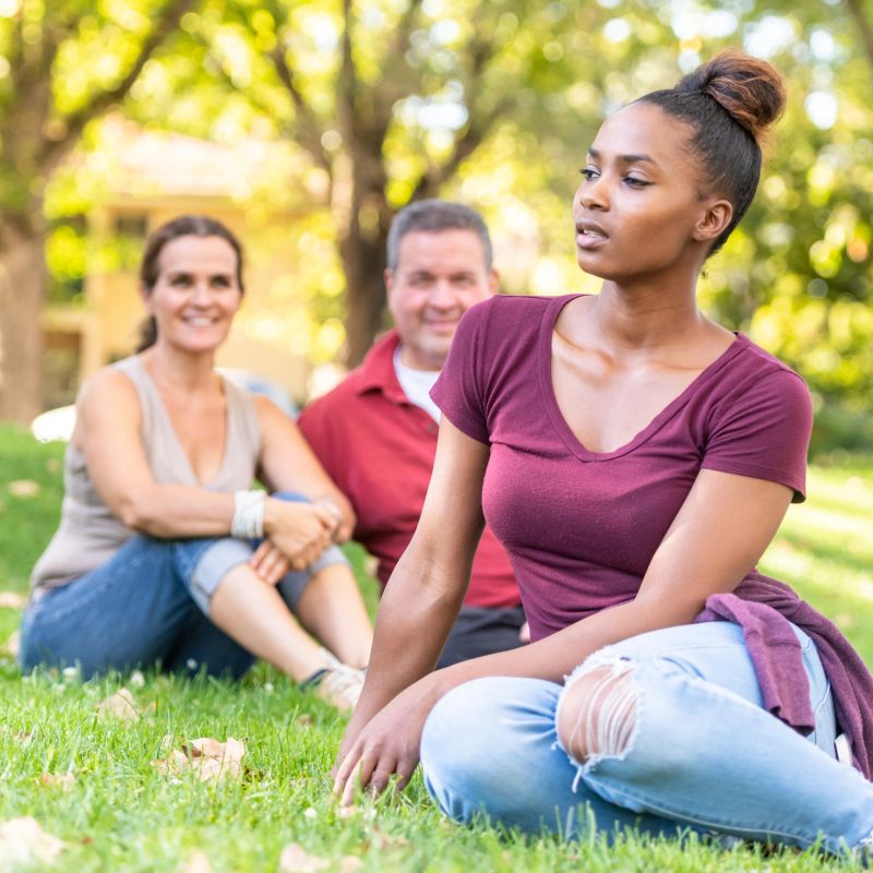 Smiling mature caucasian couple posing with their black adopted daughter sittinng on the grass at a public park