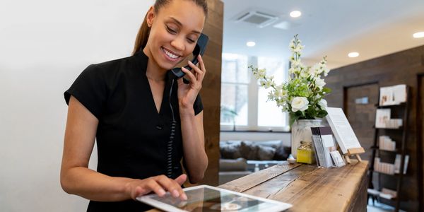 Smiling receptionist multitasking with phone and tablet at a cozy front desk.