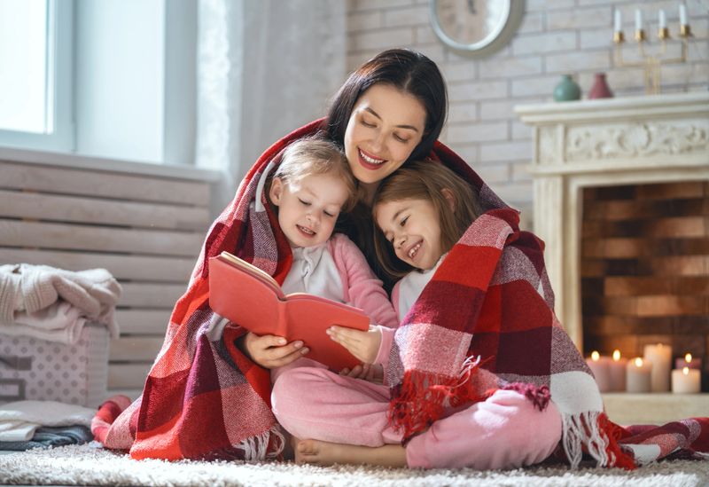 Winter portrait of happy loving family. Pretty young mother reading a book to her daughters at home.