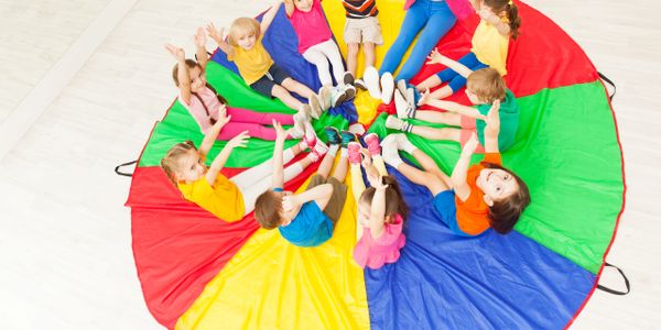 Children sitting on a colorful parachute with hands raised, smiling.