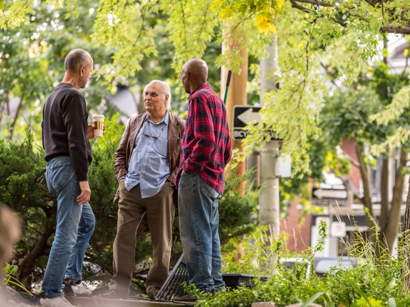 Group of senior men of various backgrounds having a friendly chat in the front yard of one man while he is raking the leafs. Bright fall scene on the road in the North American city.