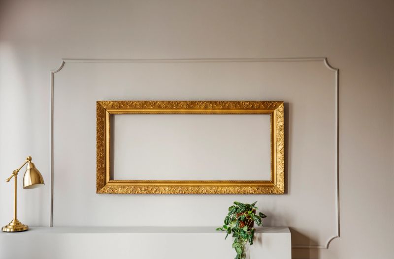 Elegant, empty golden frame inside molding on a gray wall of a stylish living room interior with lamp and a plant. Real photo.