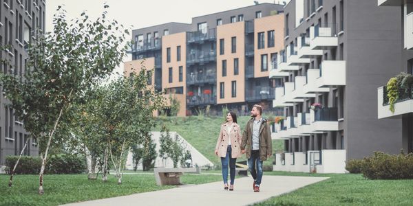 outdoor image with grass, trees, big apartment buildings and two people walking on sidewalk