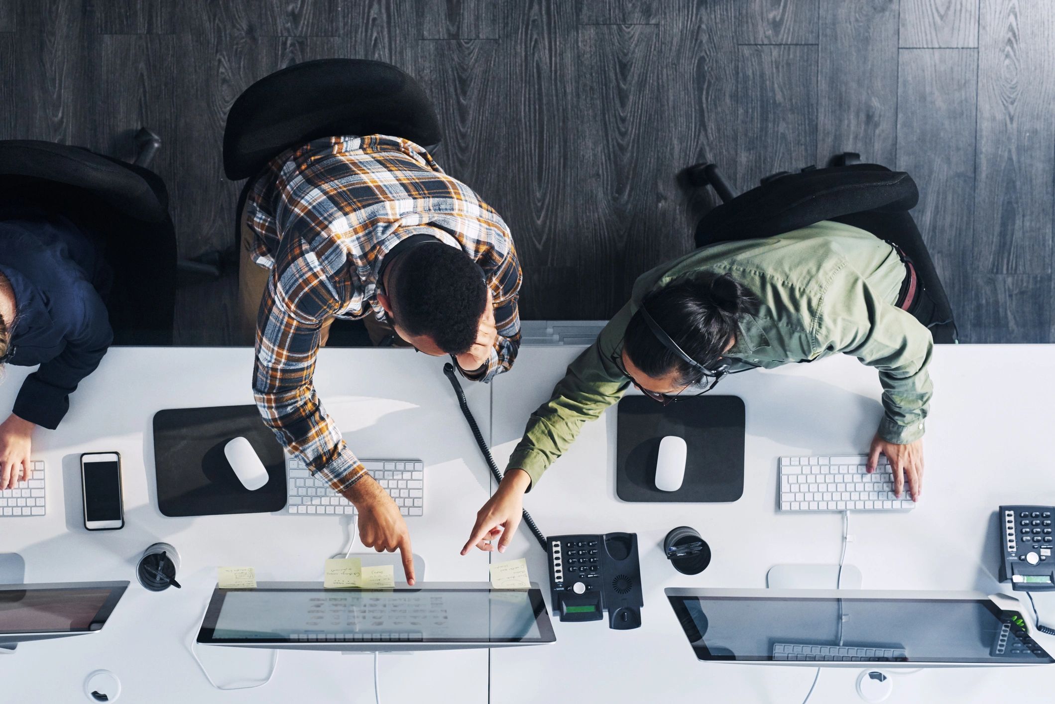 Two coworkers collaborate at a desk with computers and phones in an office setting.