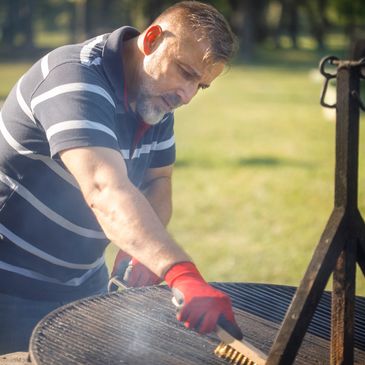 Man cleaning a barbecue grill outdoors wearing a striped shirt and red gloves.