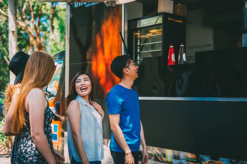 Group of young people standing in front of the food truck with delicious fast food. Diverse people buying tasty pastry, sandwiches and hamburgers in famous food truck, near the festival venue.