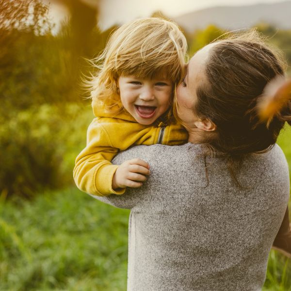 Woman holding and kissing a smiling child outdoors.