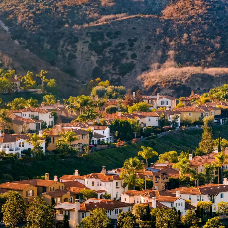 San Clemente homes on a sunny hill in California. View of beautiful homes with yards and trees on a grassy hill illuminated by sunlight. A mountain with rugged slopes can be seen in the background.