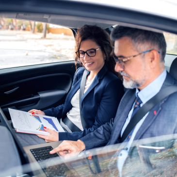 Two professionals reviewing documents and laptop in backseat of car.