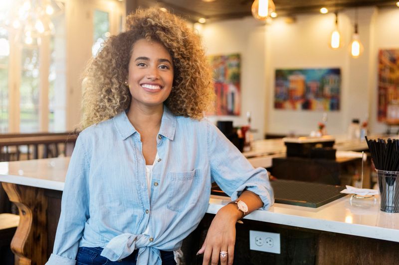 A proud young businesswoman stands in her art studio and smiles for the camera as she leans against the counter.  There are paint supplies, tables and paintings on the wall in the background.