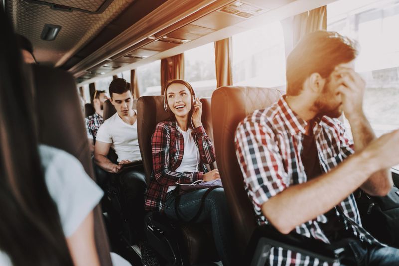 Smiling Woman Listening Music in Tourist Bus. Young Beautiful Woman Relaxing on Passenger Seat of Tour Bus in Headphones. Traveling, Tourism and People Concept. Happy Travelers on Trip