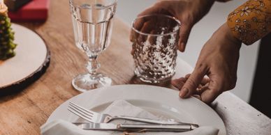 Person setting a table with glasses and cutlery on a wooden surface.