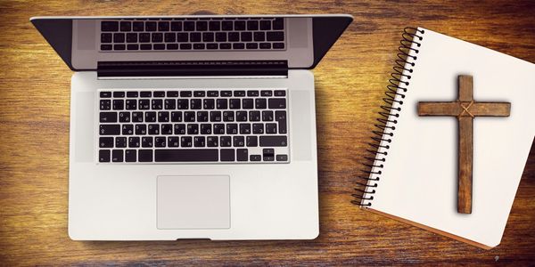 Laptop and notebook with wooden cross on wooden table.