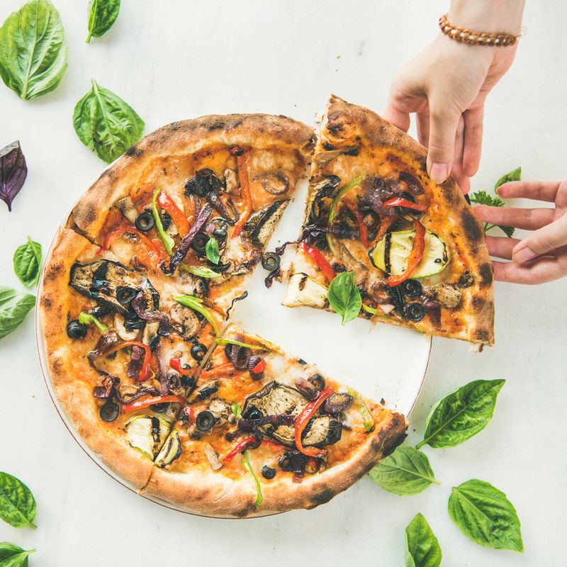 Summer dinner or lunch. Flat-lay of people's hands taking freshly baked Italian vegetarian pizza with vegetables and fresh basil over white marble table, top view, square crop