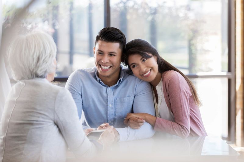 A smiling young couple embrace as they sits across a table from an unrecognizable wedding planner and discuss their upcoming nuptials.