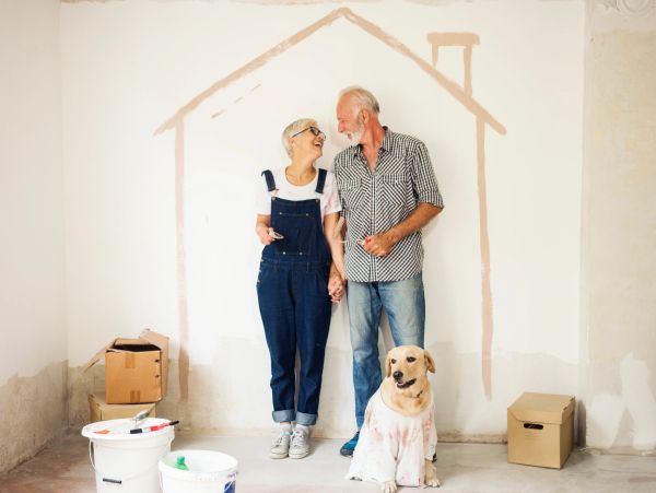 Elderly couple holding hands, smiling, with dog inside a painted house outline.