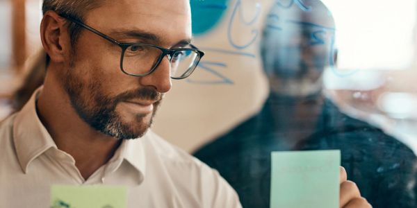 Man with glasses placing sticky notes on a glass board.