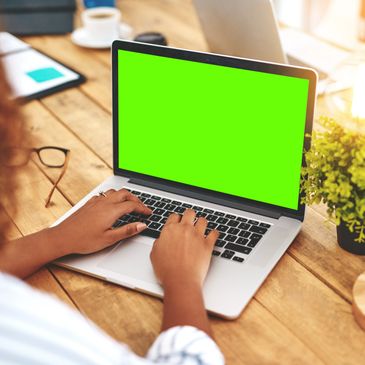 Person typing on a laptop with a green screen on a wooden desk.