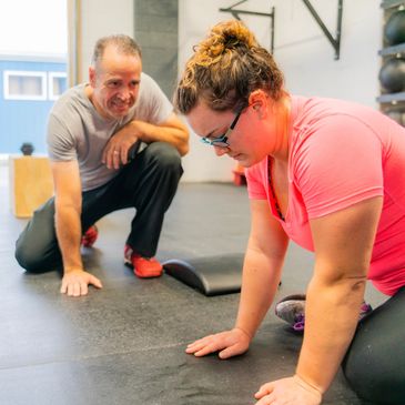 A woman in pink exercises on the floor while a man watches and supports her.