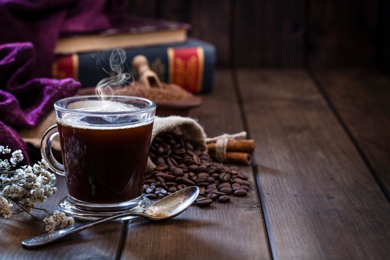 High angle view of a transparent glass coffee cup placed at the left of a rustic wooden table leaving useful copy space for text and/or logo at the center-right. An old metal spoon is in front of the coffee cup. Roasted coffee beans are behind the cup. Two old books are out of focus at background and complete the composition. Predominant color is brown. Low key DSRL studio photo taken with Canon EOS 5D Mk II and Canon EF 100mm f/2.8L Macro IS USM.