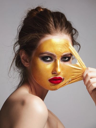 Woman peeling off a gold facial mask revealing red lipstick and smoky eye makeup.