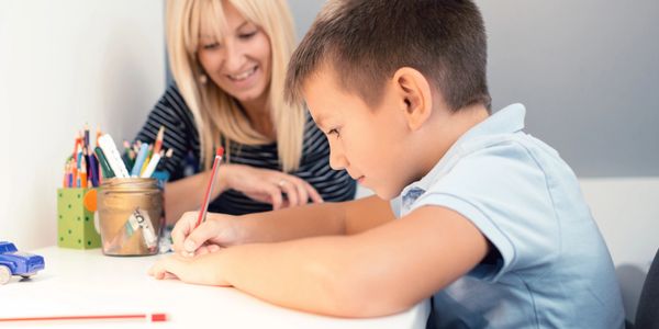 Young boy focused on writing while woman watches supportively.