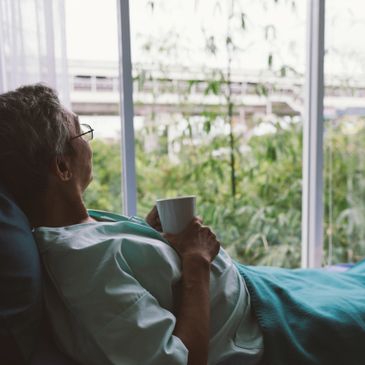Elderly person resting in bed, holding a cup and looking out the window.