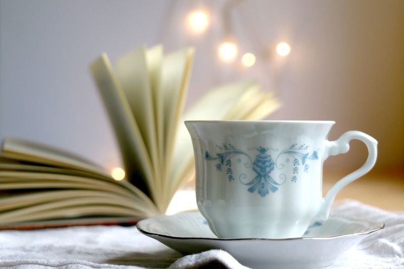 Tea in a vintage porcelain cup and open book. Fairy lights in the background. Selective focus.