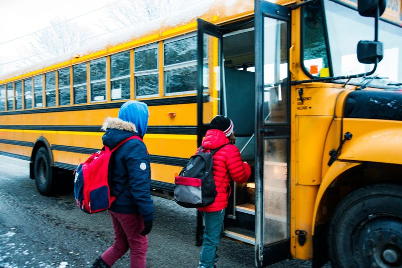 Two children getting on a school bus on a cold, snowy winter morning in the countryside.