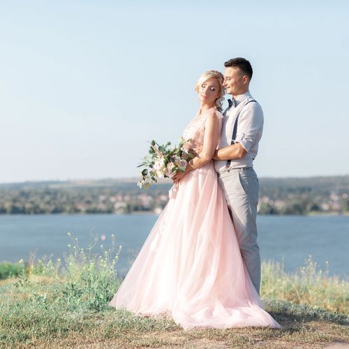 Couple in elegant attire embracing by a lakeside on a sunny day.