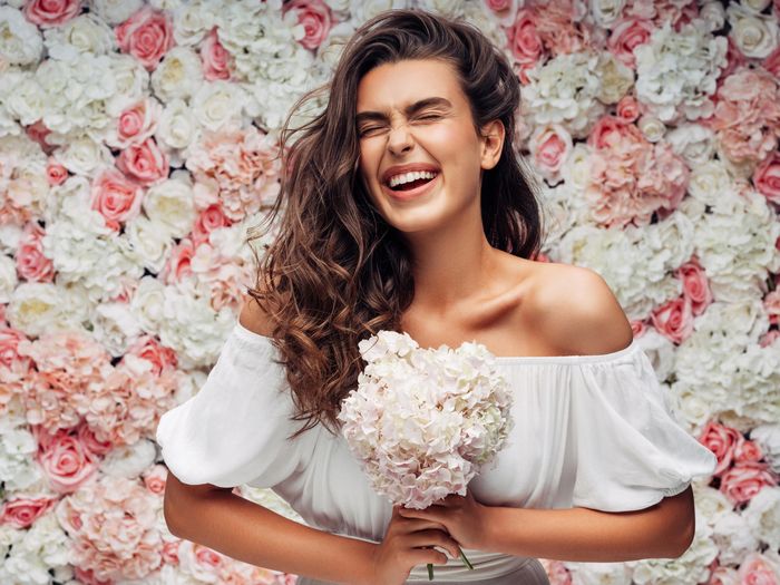 Joyful woman in white dress holding flowers against a floral backdrop.