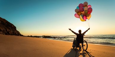 Person in wheelchair holding colorful balloons on the beach at sunset.