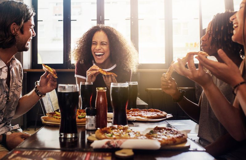 Young woman eating pizza and laughing while sitting with her friends in a restaurant. Group of friends enjoying while having food and drinks at cafe.