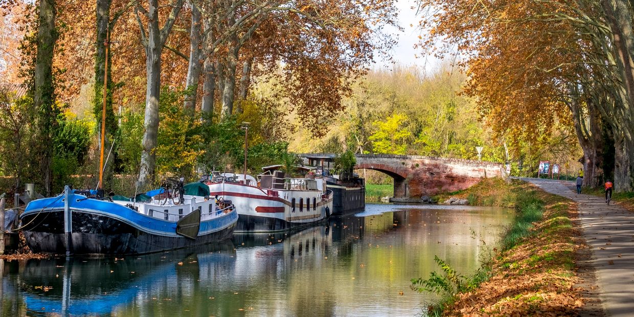 Boats docked along a tree-lined canal in autumn with a stone bridge in the background.