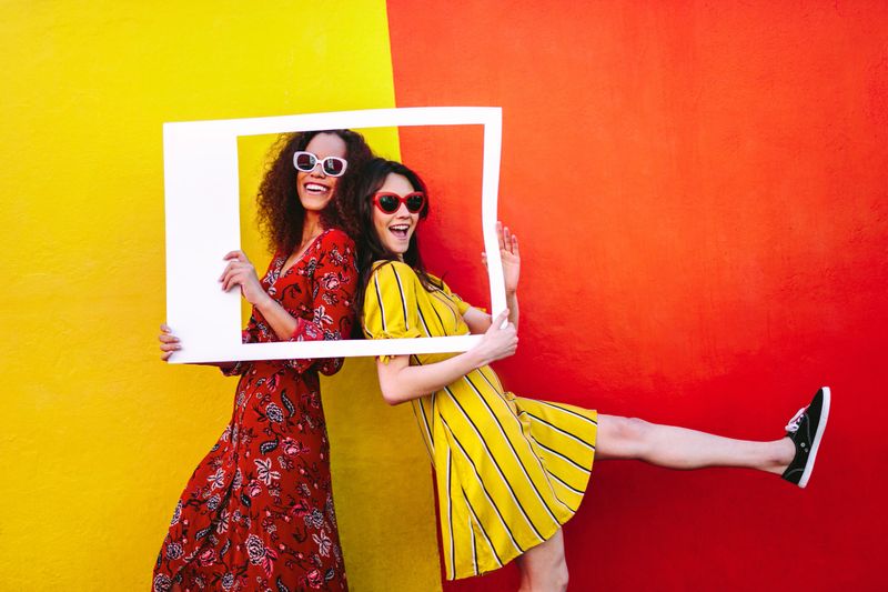 Excited girl friends with empty photo frame standing against colored wall. Women travelers wearing colorful dresses and sunglasses posing with empty picture frame.