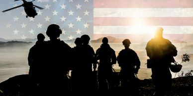 Silhouetted soldiers with a helicopter and American flag in the background at sunset.